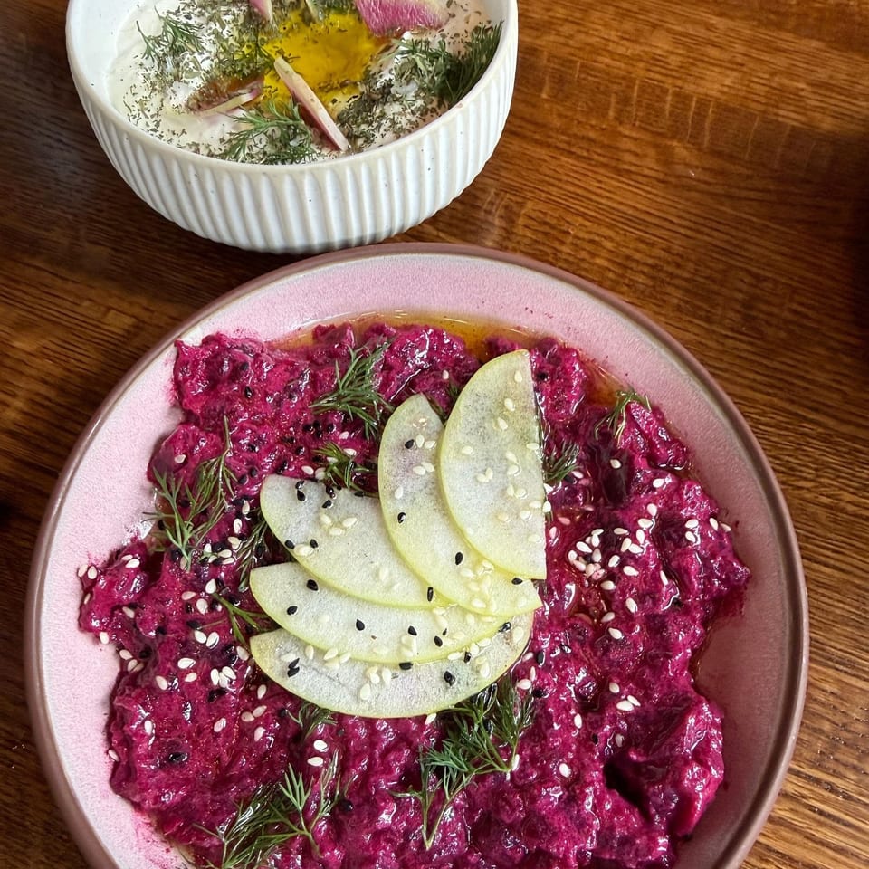 Vibrant beet salad topped with green apple slices, dill, and sesame seeds beside a herb-garnished creamy dip.