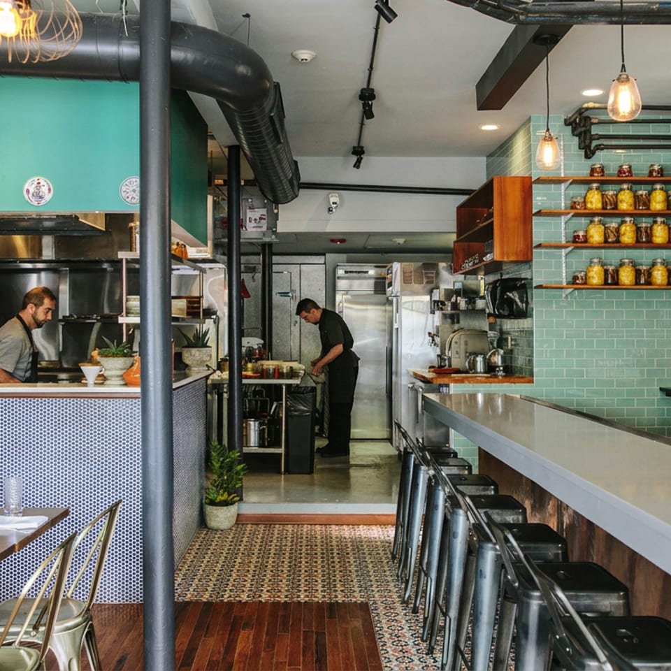 Bright restaurant interior with open kitchen, chefs working, tiled counter, bar seating, wine shelves, and set dining tables.