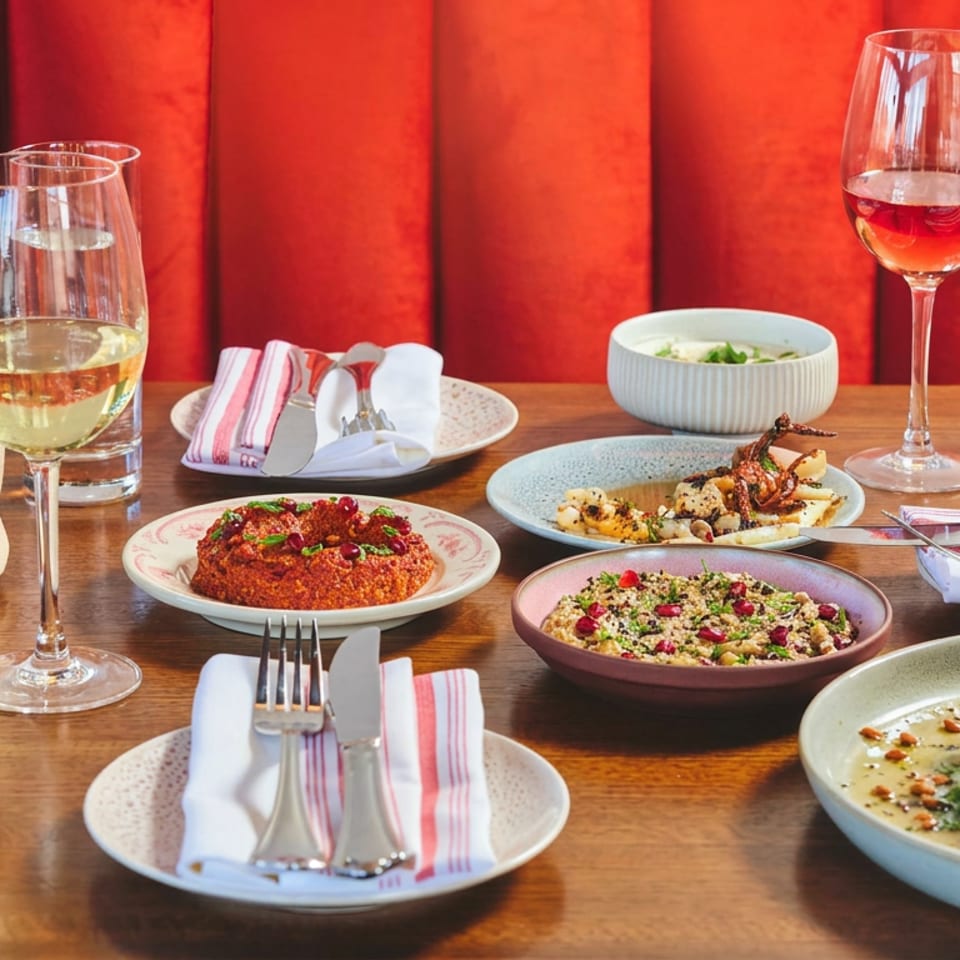 Mediterranean restaurant table spread with hummus, dips, grilled dishes, flatbread, wine, and water against a red upholstered backdrop.