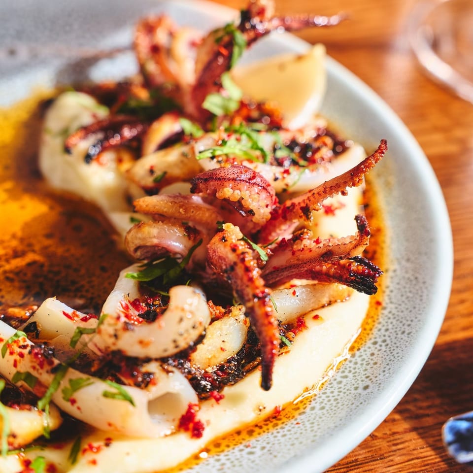 Grilled octopus tentacles with herbs and red pepper flakes arranged on a white plate atop a wooden restaurant table.
