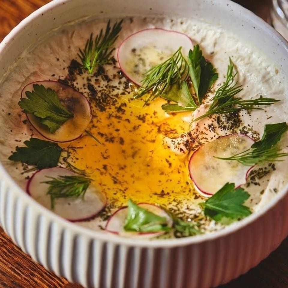 Creamy dip topped with a golden egg yolk, sliced radishes, fresh herbs, and black pepper in a white bowl.