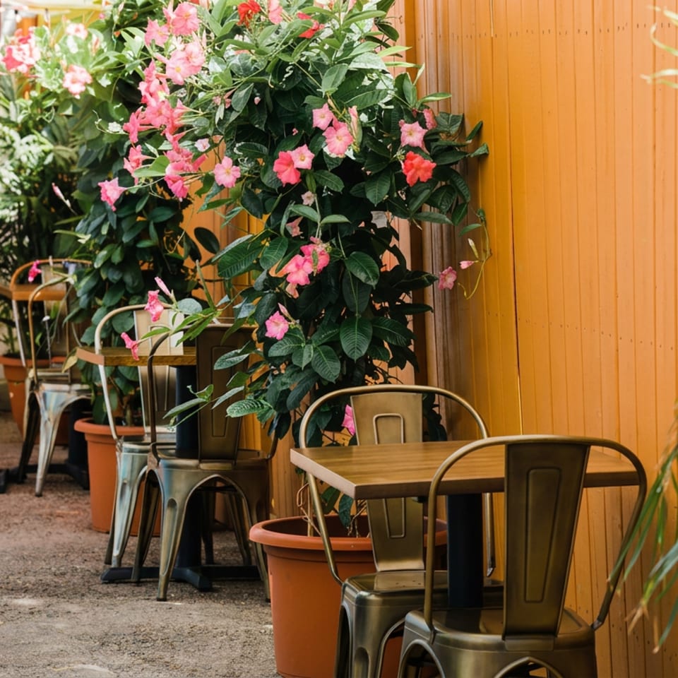 Outdoor restaurant patio with wooden tables, metal chairs, potted pink flowers, and lush greenery beside a warm wood-paneled wall.
