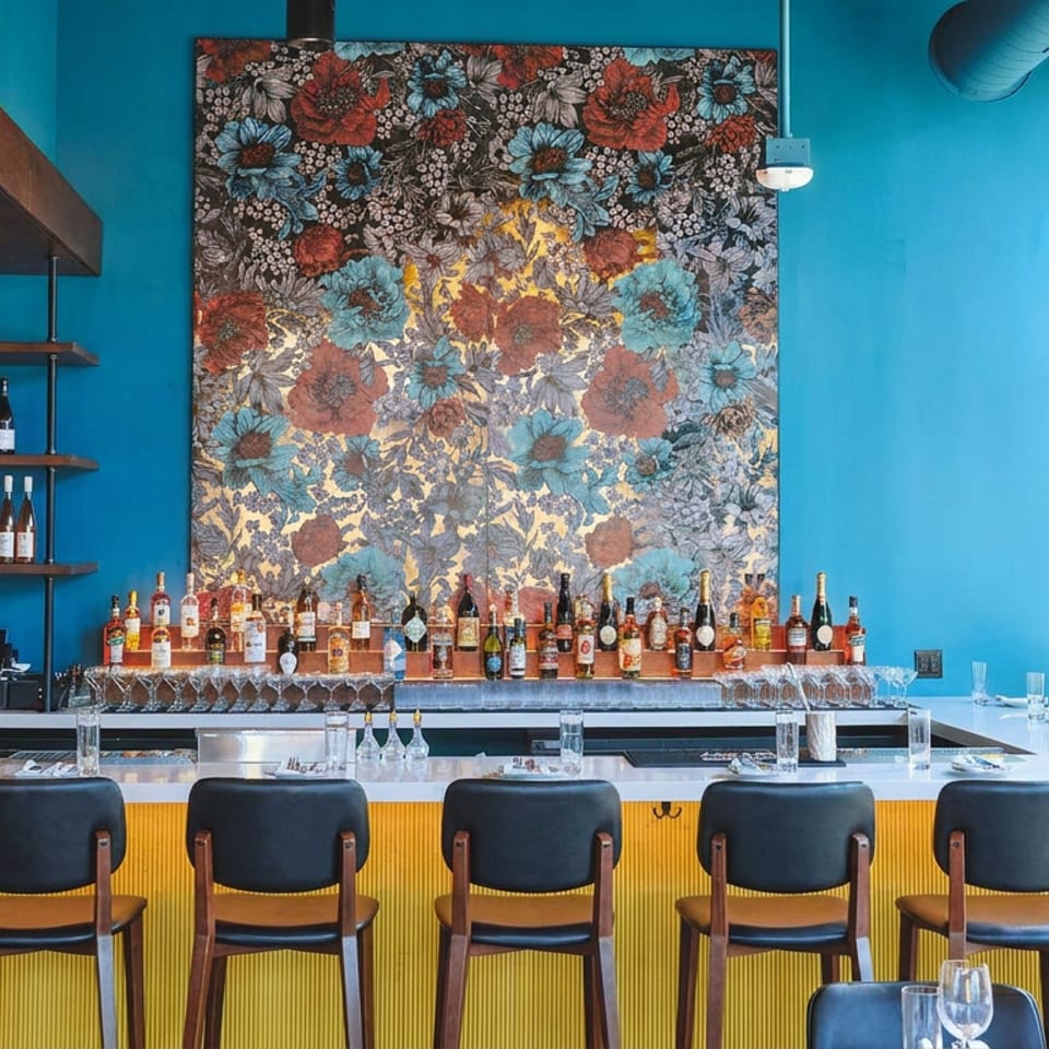 Colorful restaurant bar with liquor bottles, floral artwork, blue walls, yellow counter, black stools, and large windows.